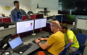 Color photo of two men wearing yellow shirts, sitting at two large computer screens to follow and control ITER plant systems that have already been commissioned. They are working on when will ITER be completed.
