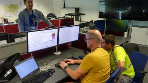 Color photo of two men wearing yellow shirts, sitting at two large computer screens to follow and control ITER plant systems that have already been commissioned. They are working on when will ITER be completed.