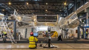 Photo de deux hommes de metrologie qui portant des casques bleus et des gilets de sécurité jaunes, debout parmi de grandes poutres en acier. Ils effectuent des mesures précises pour garantir les tolérances rigoureuses requises dans l'assemblage de la machine ITER.