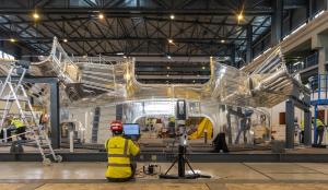 Photo de deux hommes de metrologie qui portant des casques bleus et des gilets de sécurité jaunes, debout parmi de grandes poutres en acier. Ils effectuent des mesures précises pour garantir les tolérances rigoureuses requises dans l'assemblage de la machine ITER.